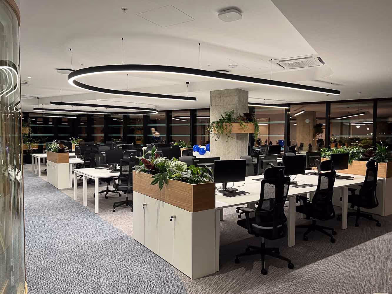 A modern, open-plan office with several white desks and black chairs. The ceiling features curved, contemporary lighting. Each desk has computer monitors and plants. Large windows in the background provide natural light.