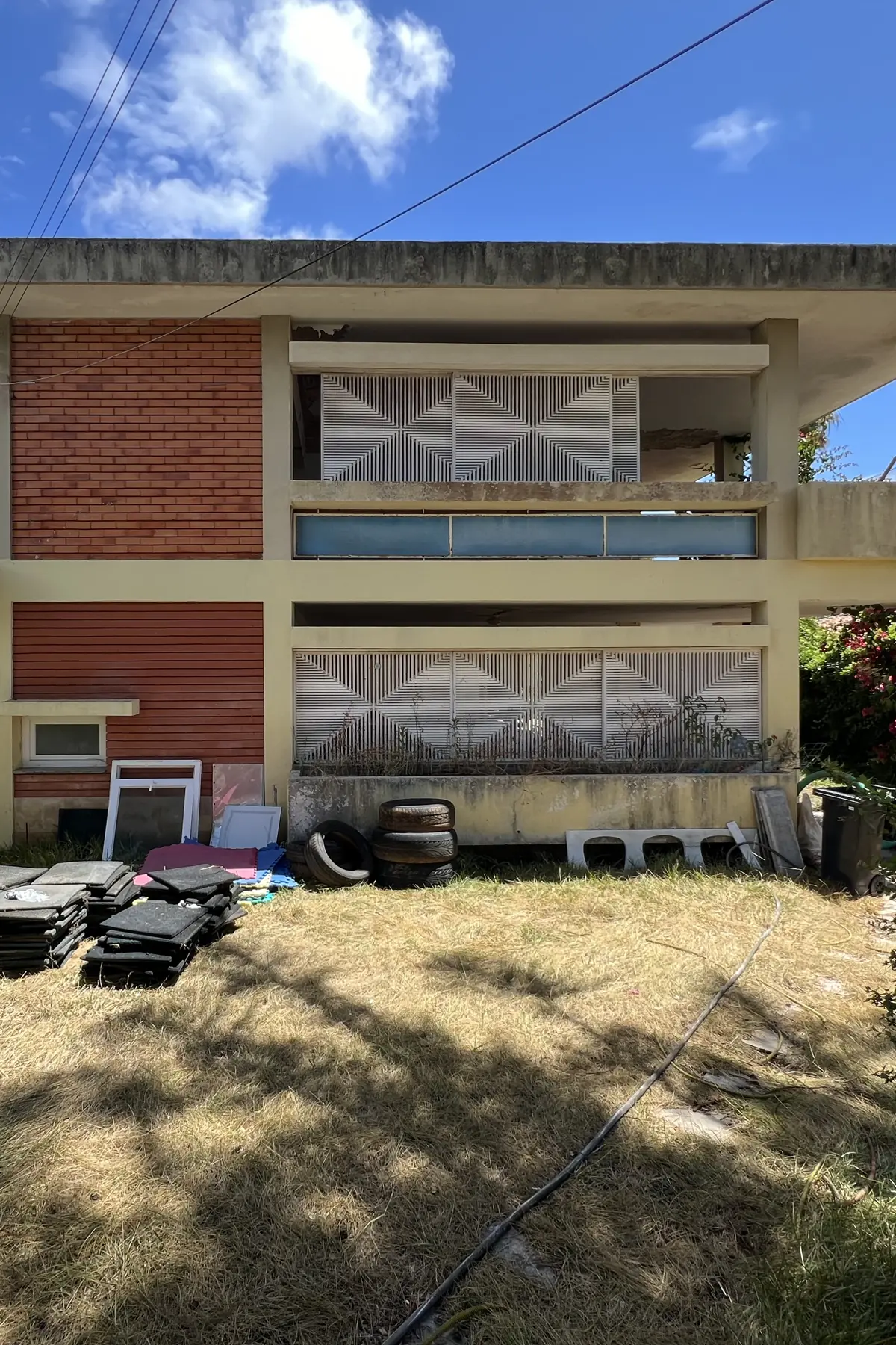 The garden elevation square on: a brick panel to the left, two recessed floors with white screen panels and a blue band, the roof slab projecting above, and materials stacked against the base wall.