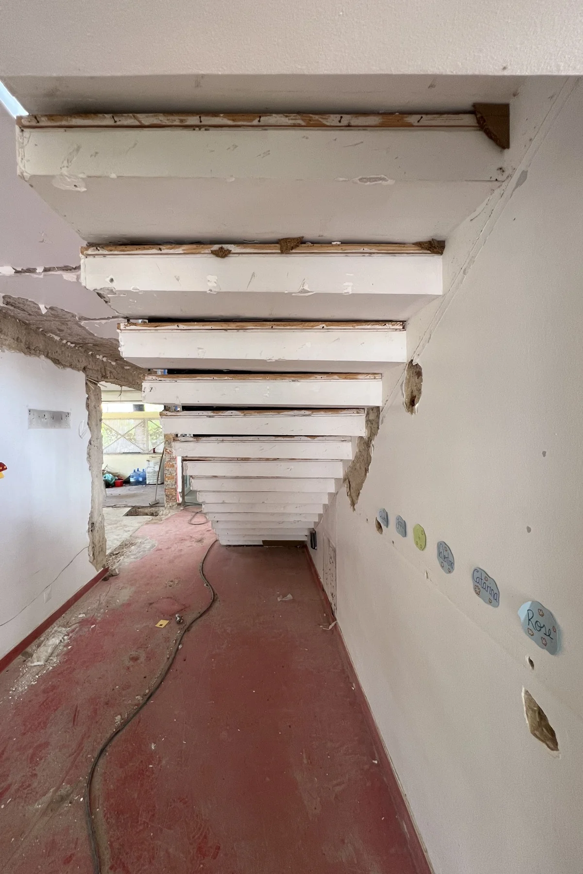 A corridor running under the stair: the undersides of the steps step down across the ceiling one after another, a red floor below, and a cable run along it.