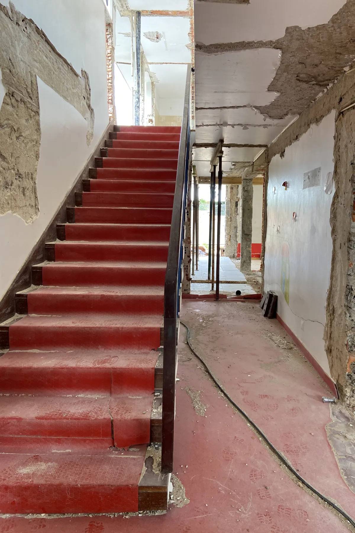 A straight flight of stairs finished in red, rising against a wall with its finish removed, with a dark metal handrail and, beyond it, a corridor with uprights.