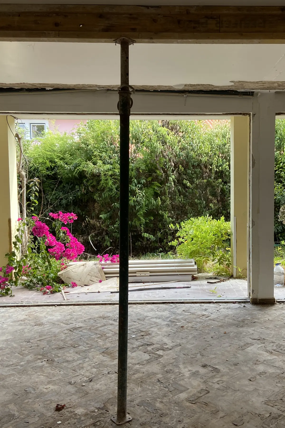 A ground-floor room with its partitions removed, a metal upright under the beam, and the wall open onto the garden, with bougainvillea in flower against the hedge.