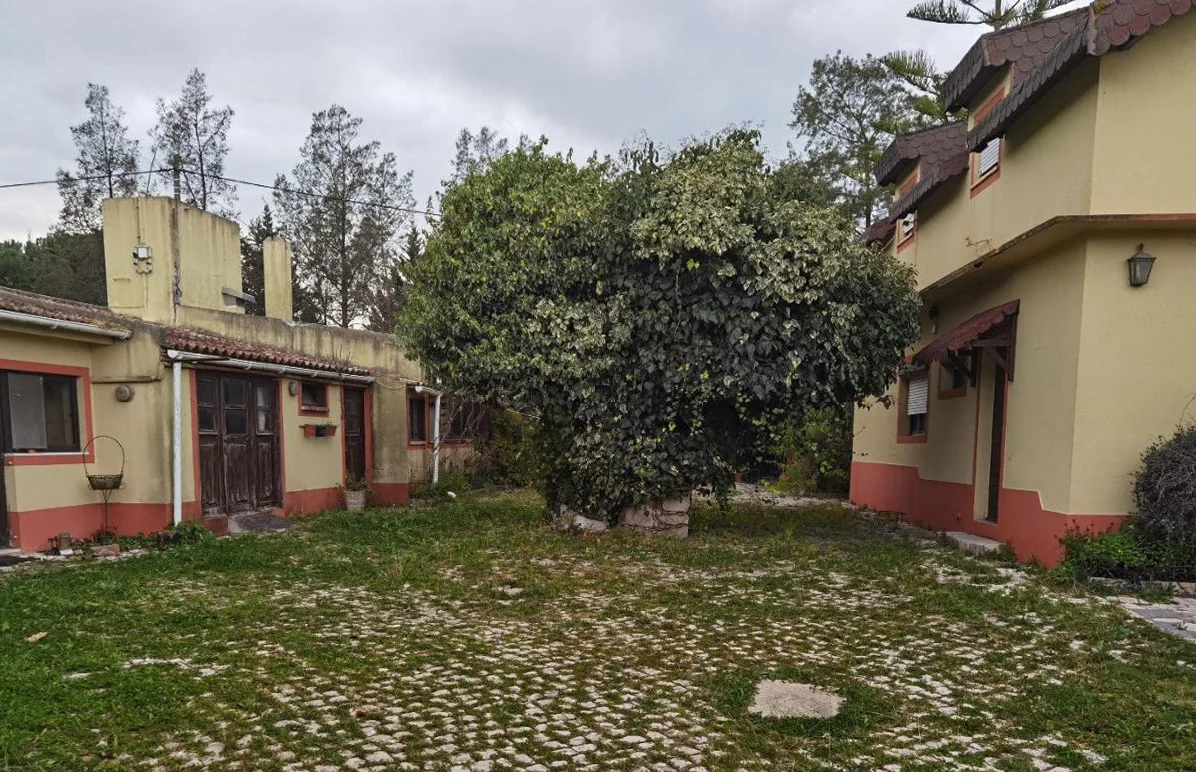 A courtyard between single-storey buildings rendered in pale yellow with red-painted bases, an overgrown lawn and a cobbled path, and a taller house with a tiled roof at the far end.