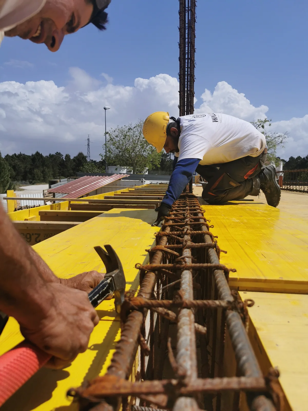 Hands tying reinforcement bars over yellow formwork, with a worker in a hard hat beyond.