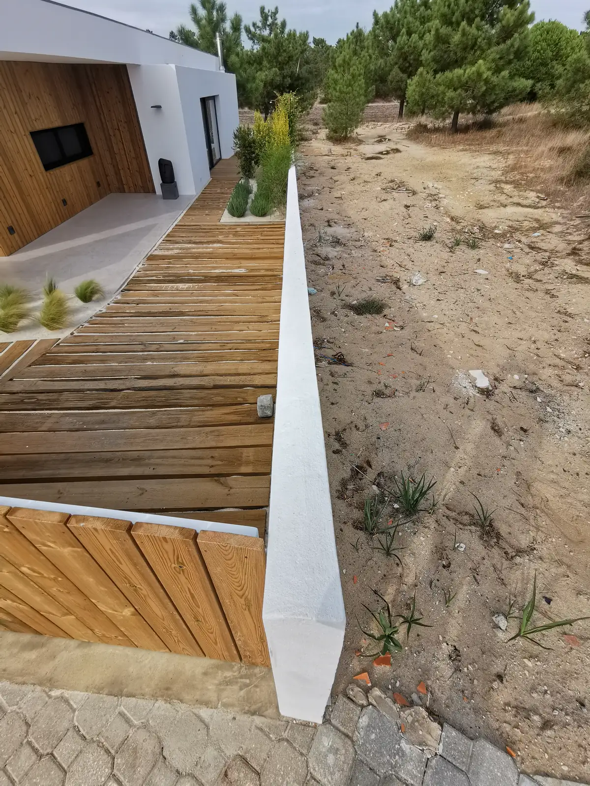 A timber deck running along a white rendered wall, with sandy ground and new planting beside it.