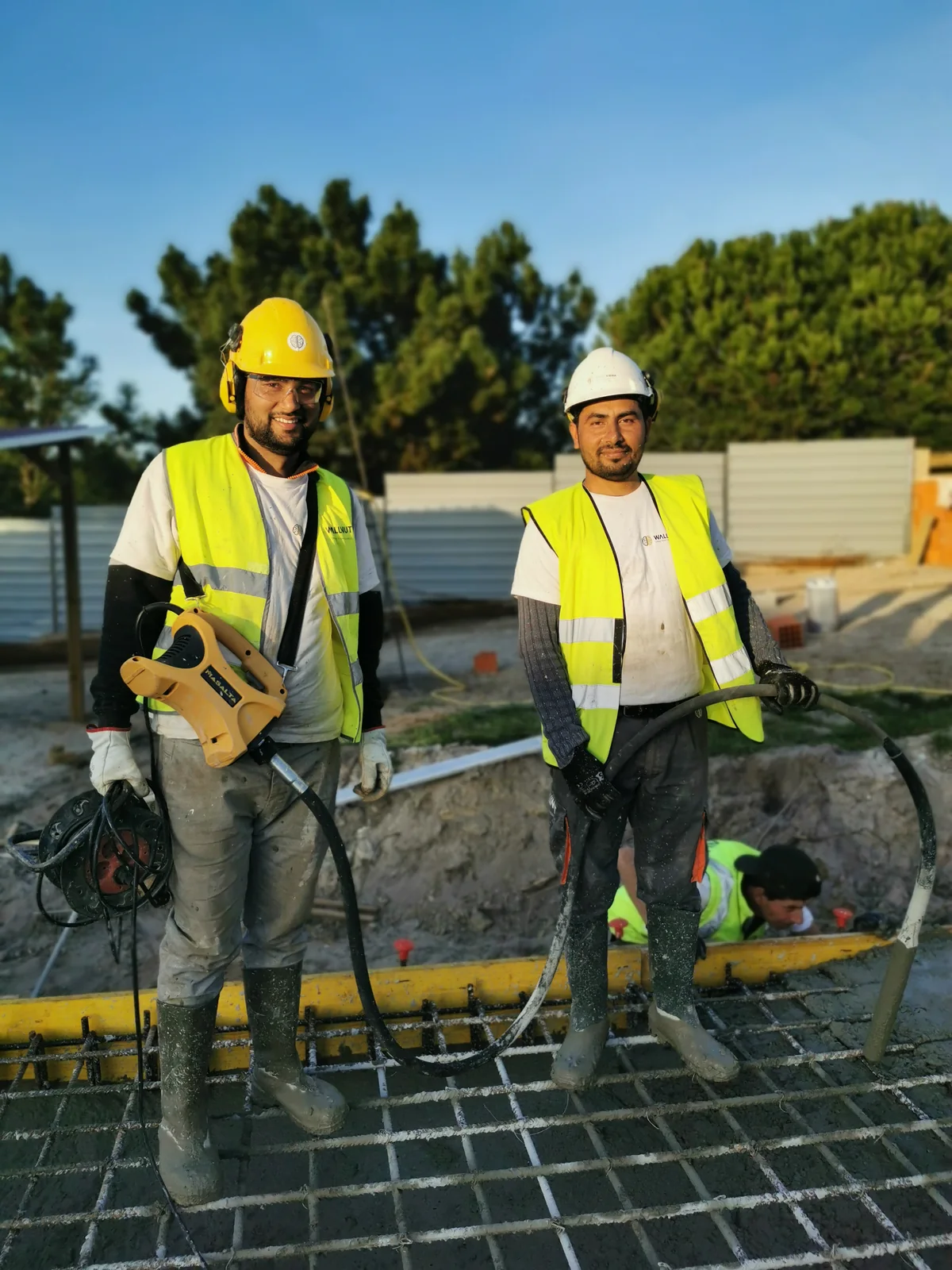 Two workers in hard hats and high-visibility vests standing on a reinforcement mesh, with pine trees behind them.