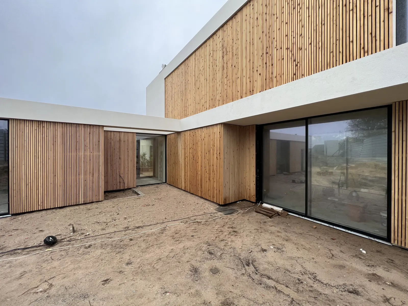 A courtyard between a white rendered volume and a wall of vertical timber cladding, with large sliding glazing and sand underfoot.
