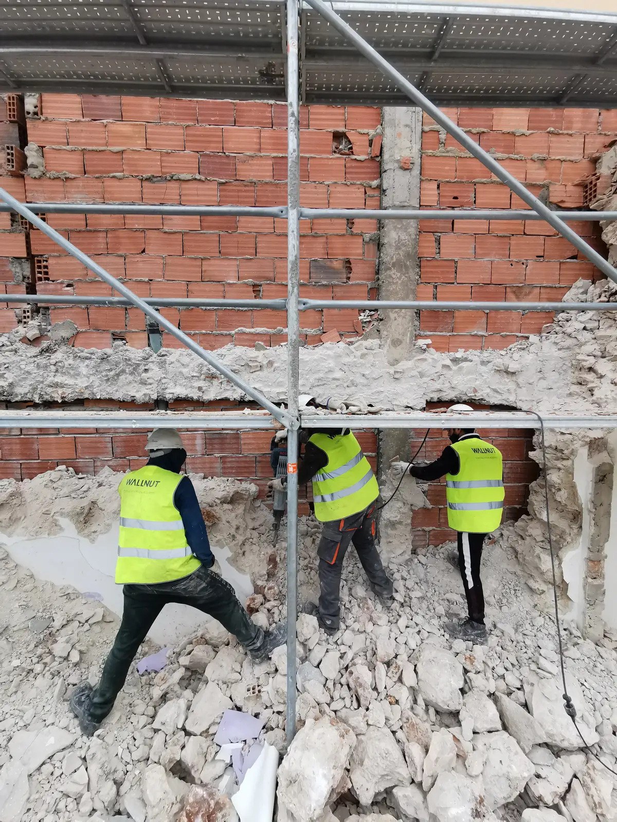 Three workers in high-visibility vests standing on rubble in front of a brick wall being taken down, with scaffolding above.