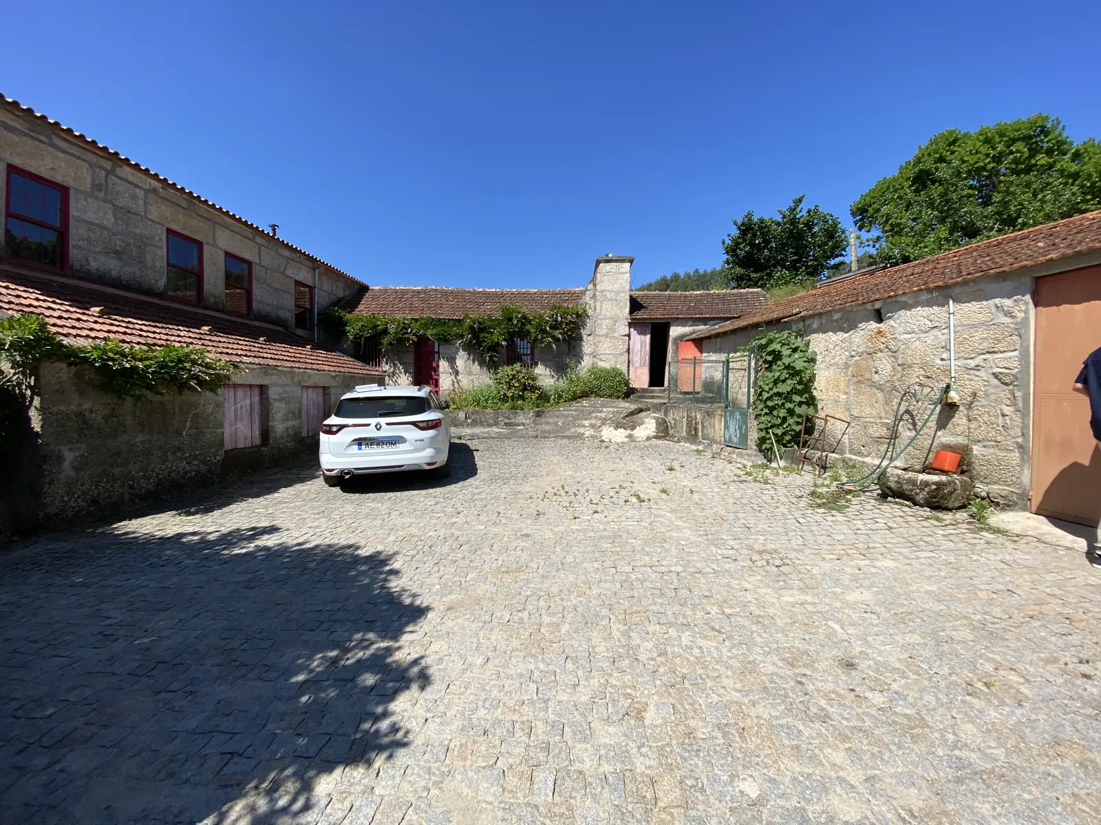 The courtyard as found: granite buildings with terracotta roofs around a cobbled yard, a car parked on it, dry stone walls and mature trees behind.