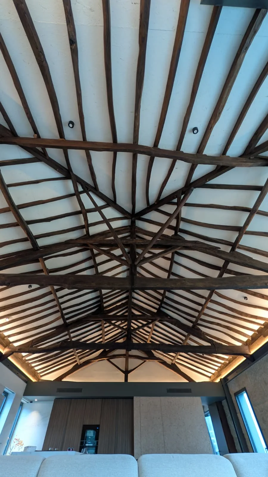The restored timber roof structure of the manor seen from directly below: dark beams radiating from a central hip over pale infill panels, with concealed cove lighting at the wall head.