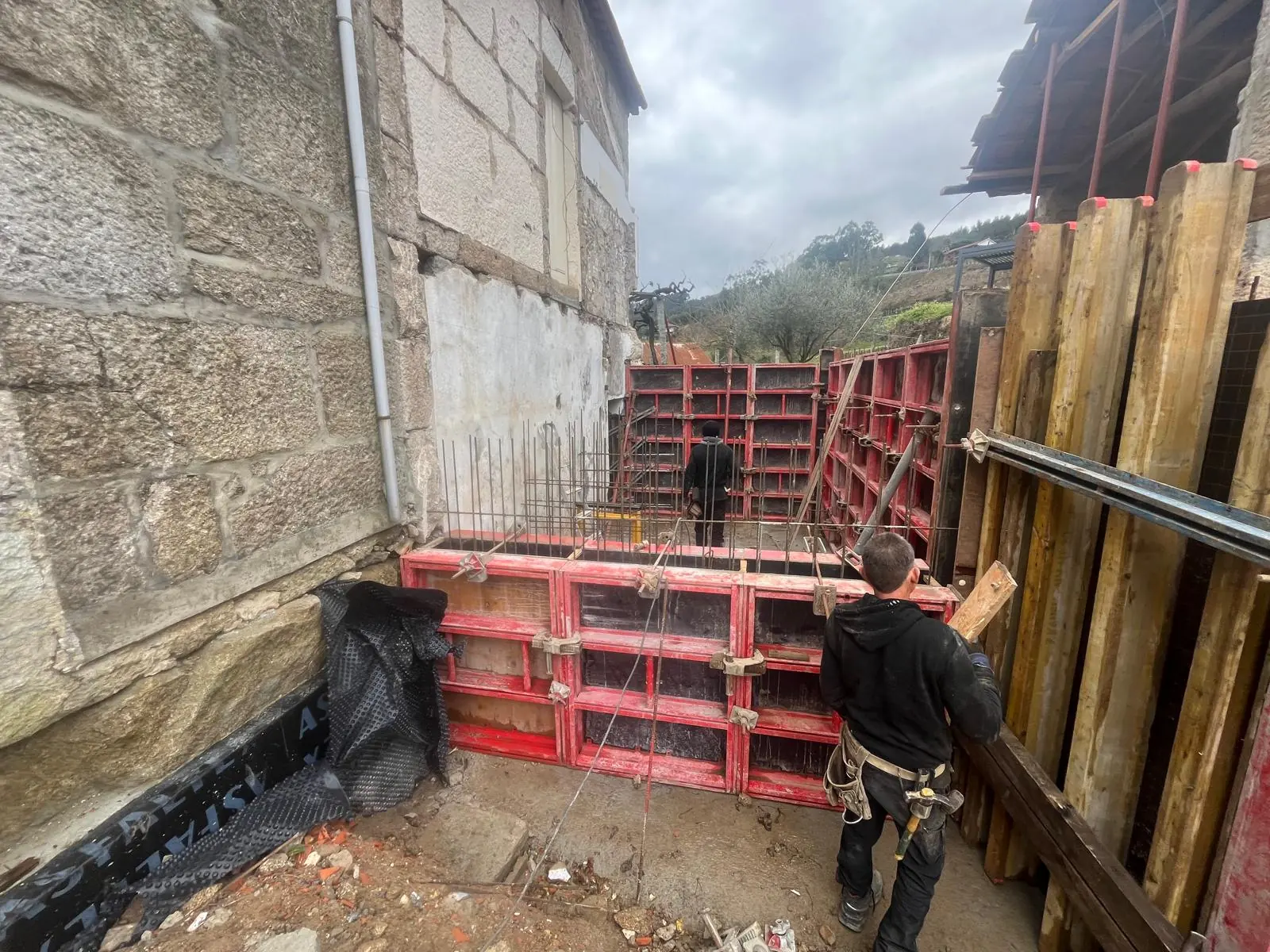 Red formwork panels set against the outside of the granite wall for a new concrete element, waterproofing at the base and a worker adjusting a prop.