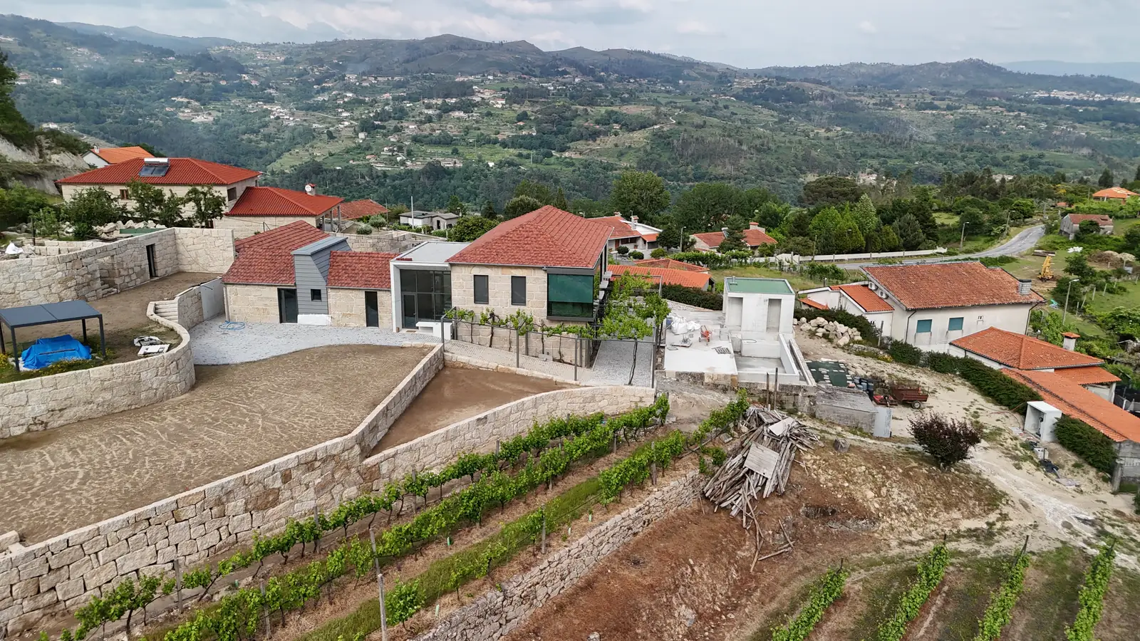 The same estate five years later: the granite buildings re-roofed, a new dark glazed support building beside them, new dry-stone retaining walls stepping down through replanted vines, and the valley opening out behind.