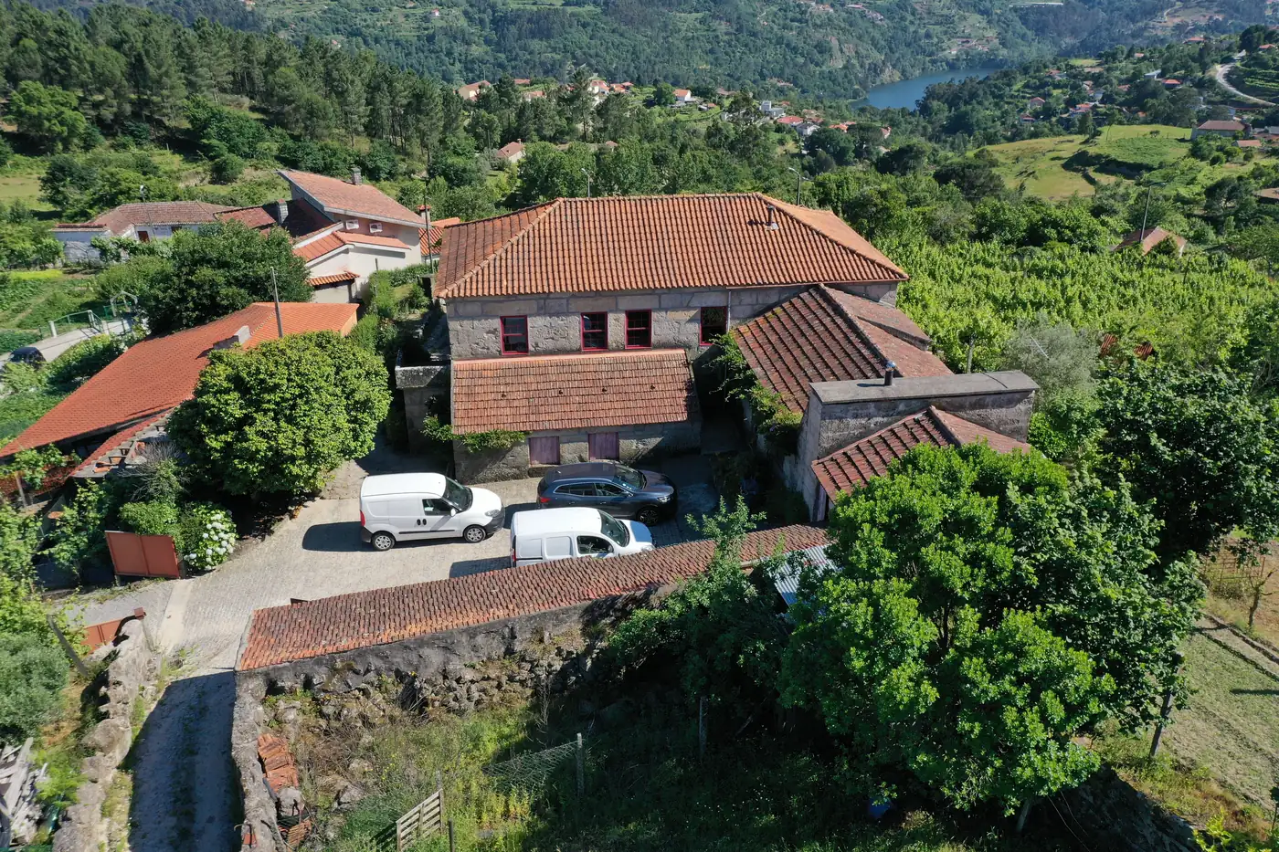 The estate from the air in summer: the granite manor with its terracotta roofs around a courtyard, dry-stone walls stepping down through vines and orchard, and the Douro visible in the valley beyond.