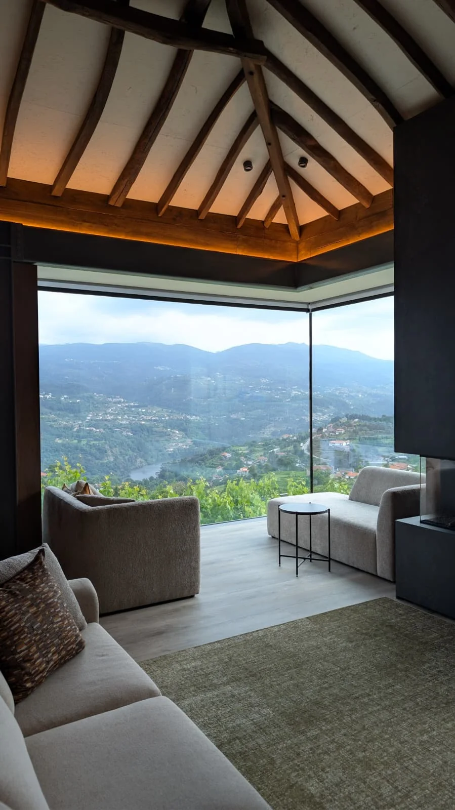 A finished sitting room under the timber roof, with a corner of full-height glazing onto the valley, low sofas, a round side table and a green rug.