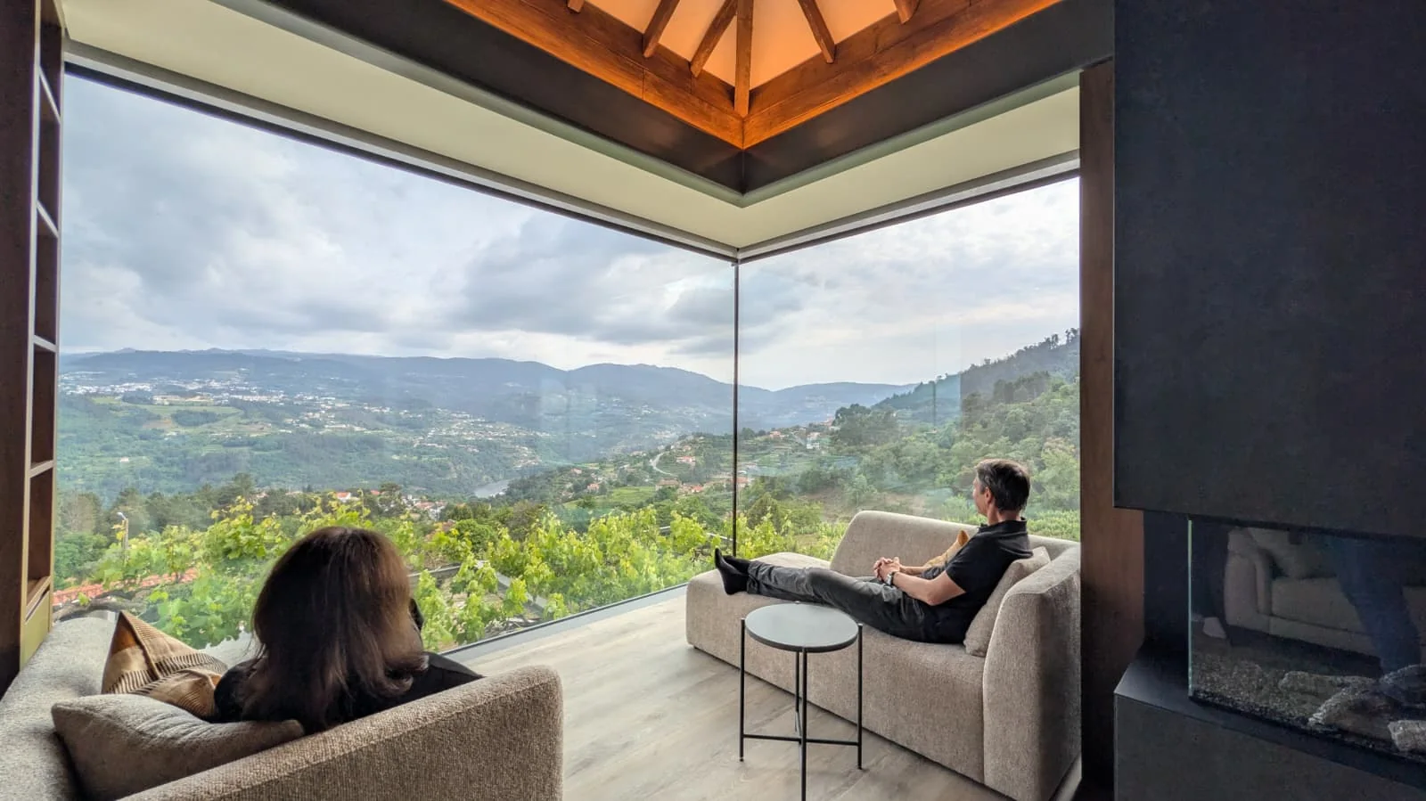 The finished sitting room: a corner opened in full-height glass with two people seated looking out over vineyard terraces and the hills beyond, a timber ceiling above and a dark fireplace wall to the right.