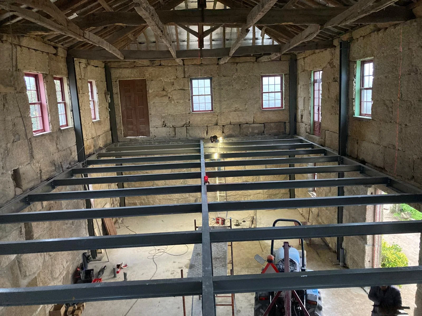 The same kind of space during the work: a grid of steel joists spanning between the granite walls at first-floor level, the old roof timbers still overhead and two workers below.