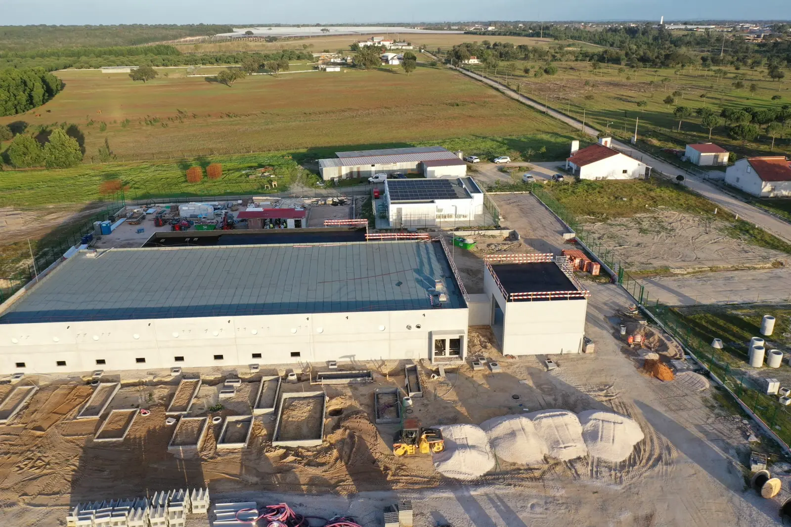 An aerial view of a long white production building with a metal roof, complete, with a second smaller building beside it and foundations still being worked in the foreground.