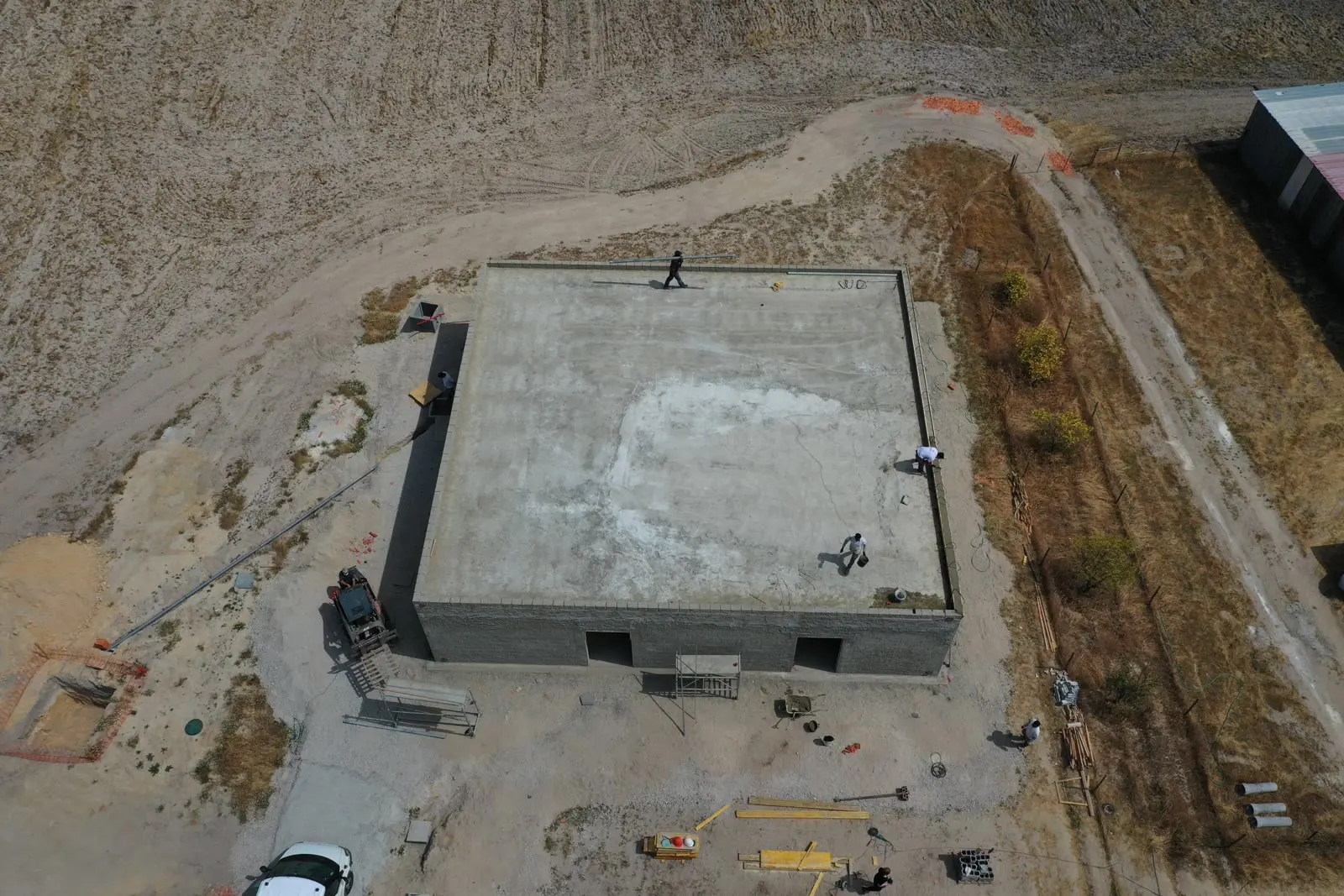 An aerial view of a single concrete building shell with its roof slab poured, standing alone on a cleared platform with plant and materials around it.