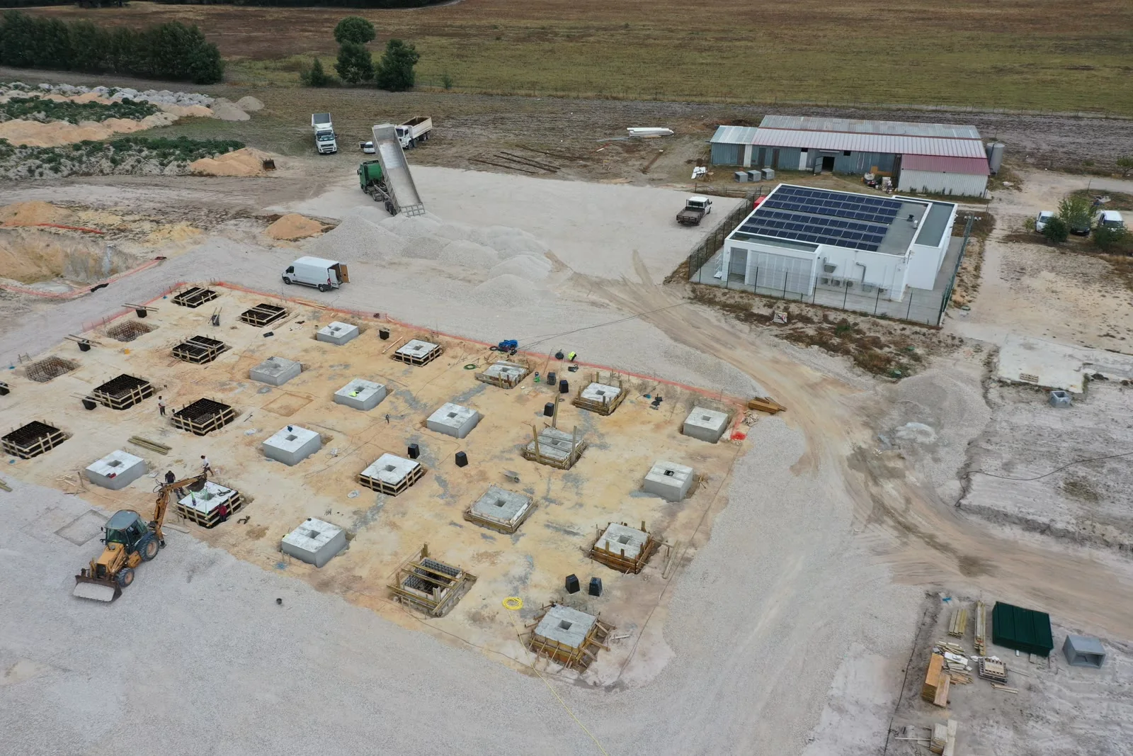 An aerial view showing a completed white building with rooftop solar panels beside a grid of freshly cast pad foundations laid out across a cleared platform.