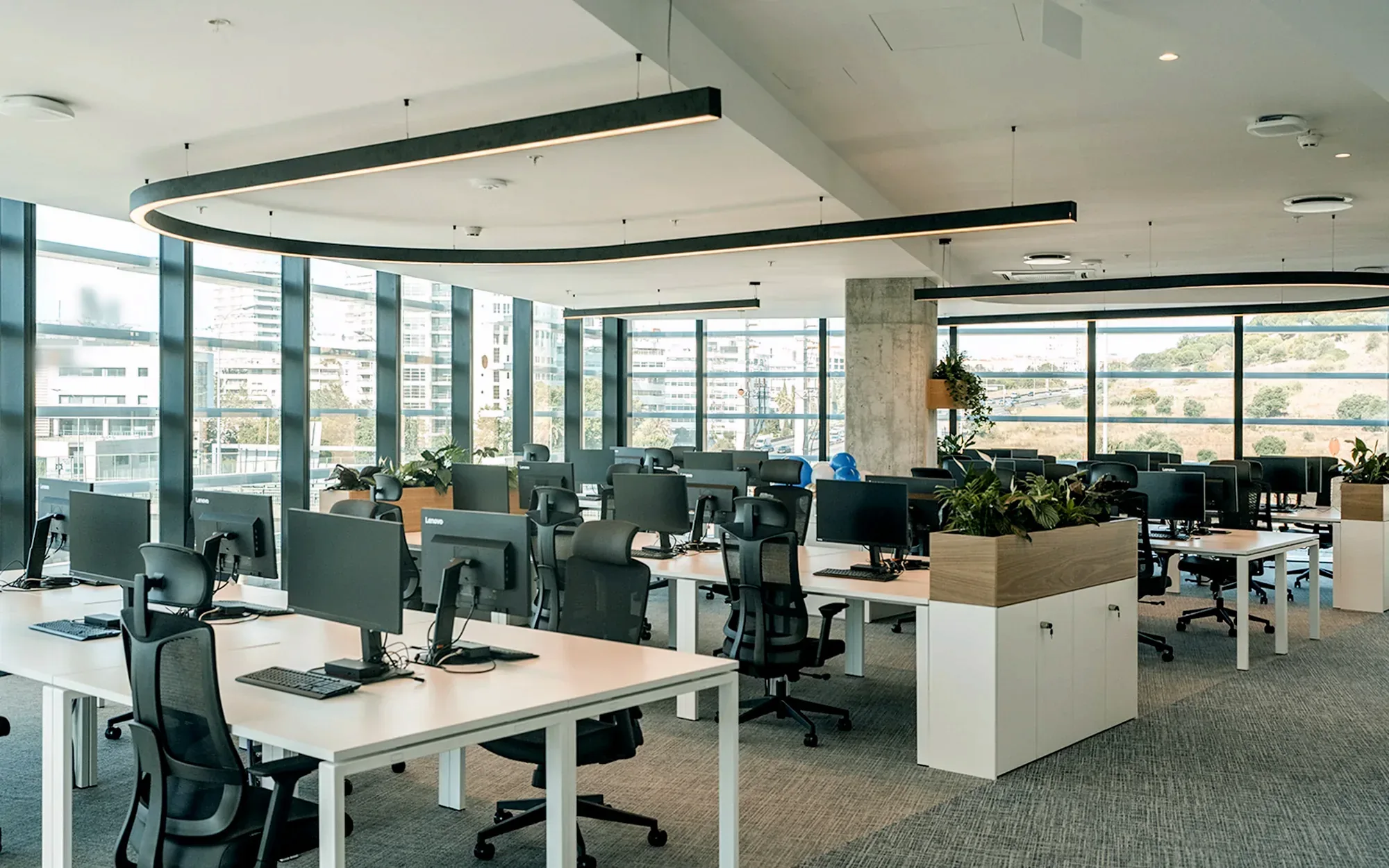 Open workspace with white bench desks, black mesh task chairs and low white cabinets topped with light timber planters, against a two-sided glazed façade looking out to a wooded slope.