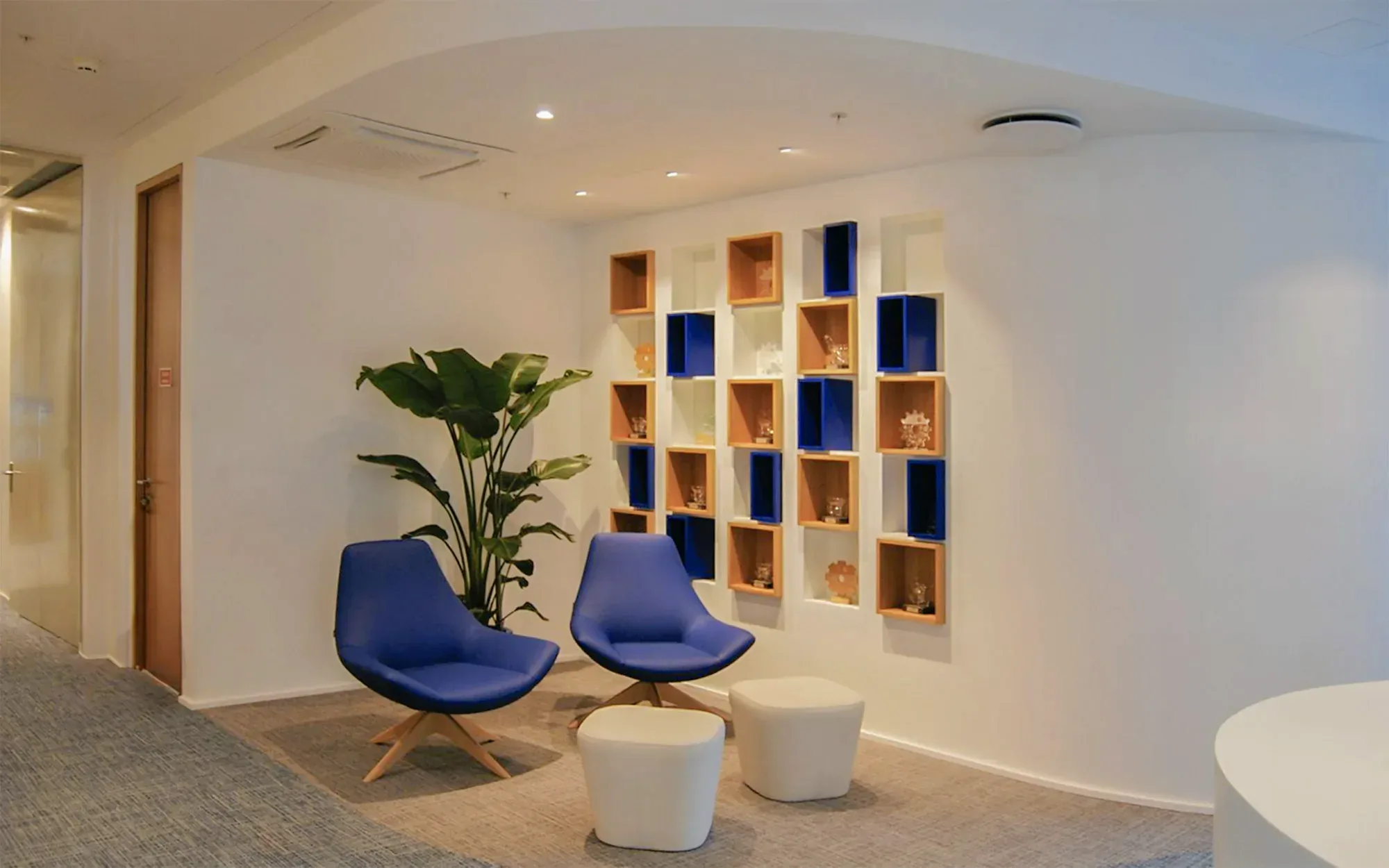 Waiting area with blue swivel armchairs and white poufs beside a white wall of square shelf boxes, some in light timber and some lacquered dark blue, under a ceiling that curves down to meet it.