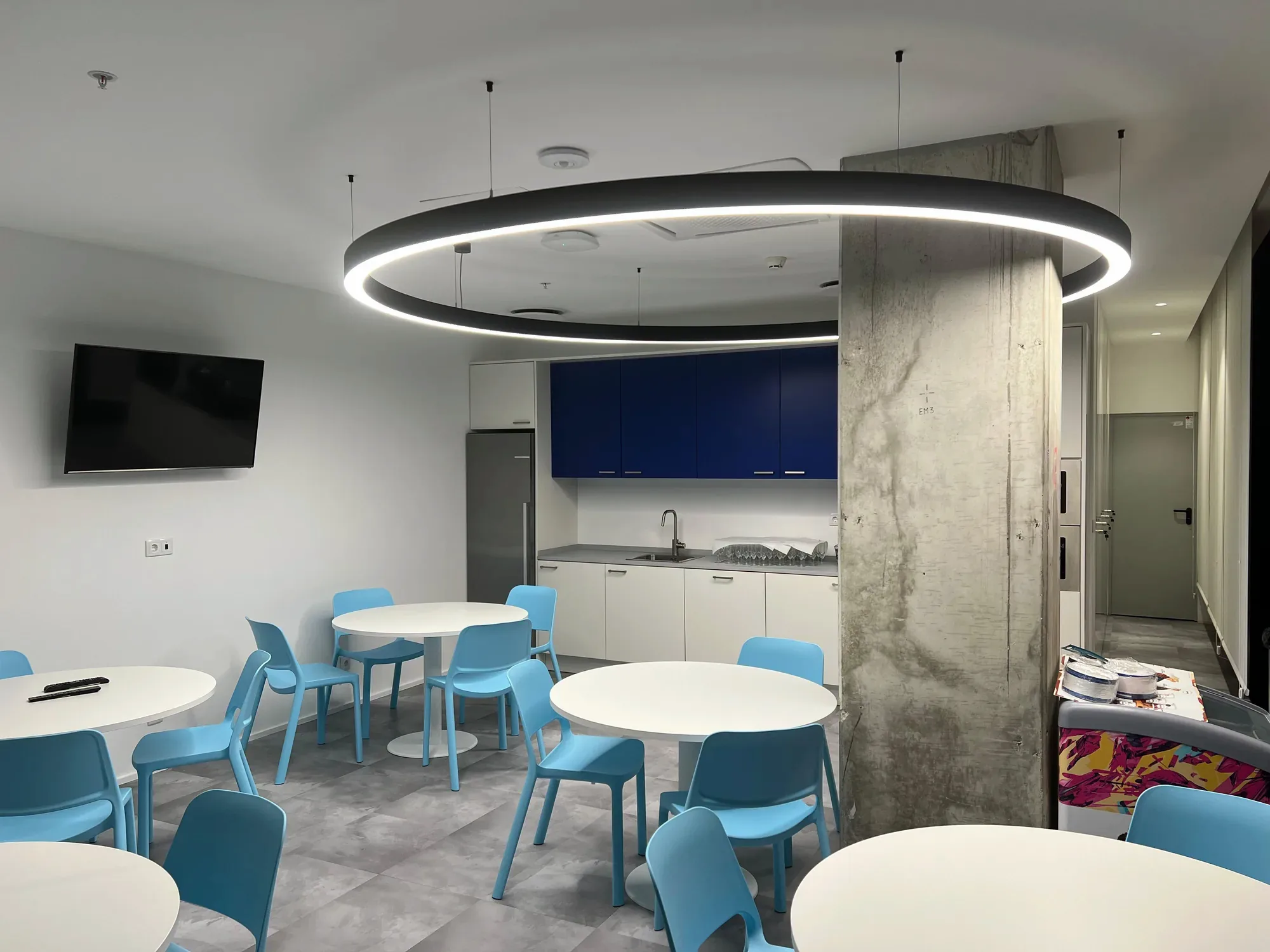 An eating room with round white tables and pale blue moulded chairs on grey tiled flooring, a white and dark blue kitchenette along the back wall, and a large black ring pendant overhead beside an exposed concrete column.