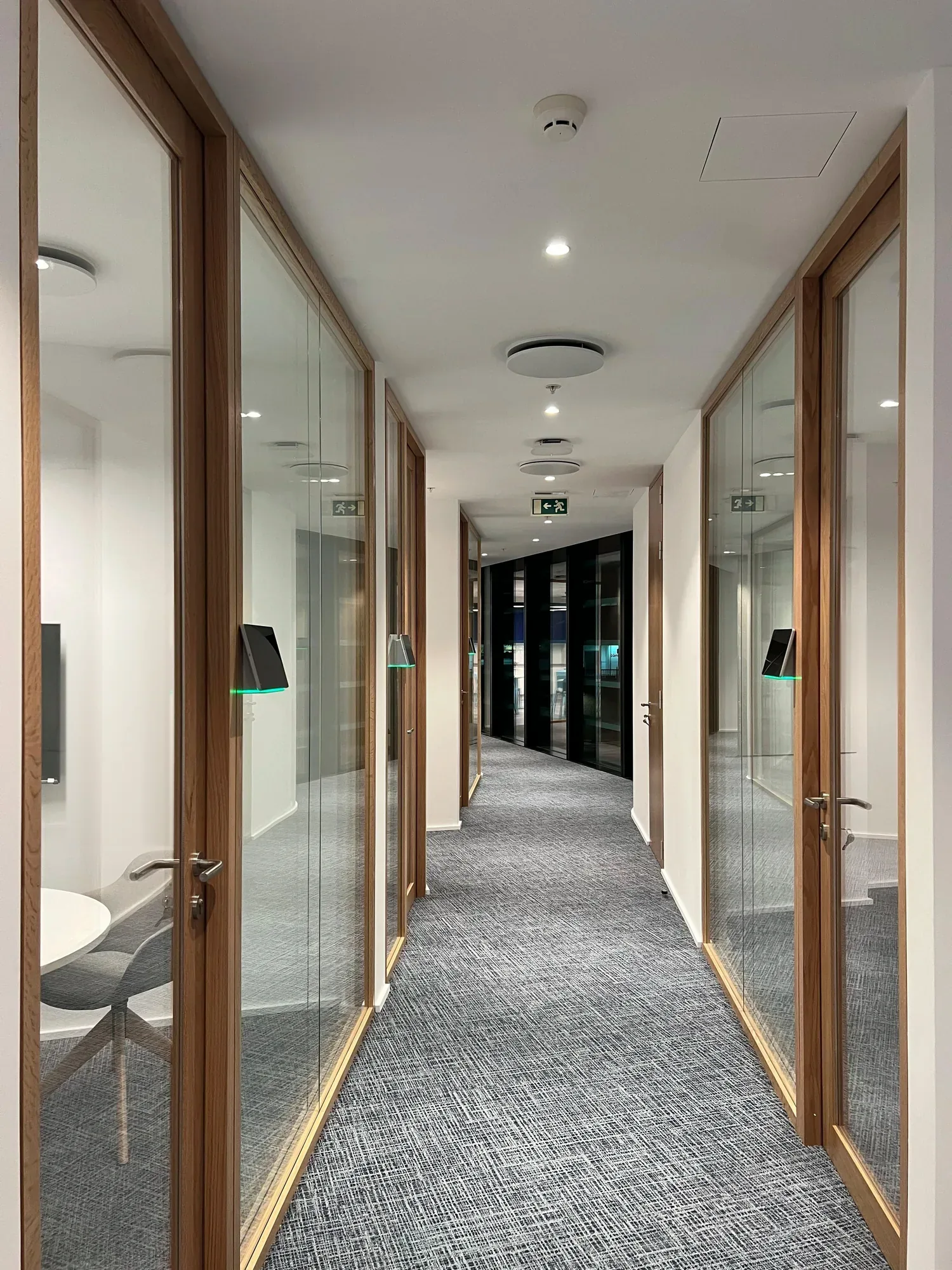Corridor between two rows of meeting rooms in oak-framed clear glass, with a lit room-booking panel beside each door, grey textured carpet, and a dark glazed wall closing the view at the far end.