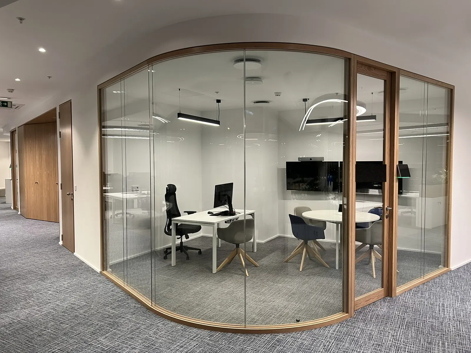 A meeting room enclosed in a single curved glass wall framed in light oak, its whole plan visible from outside: a desk, a screen, a round table and four chairs, with a linear pendant inside and a ring pendant beyond.