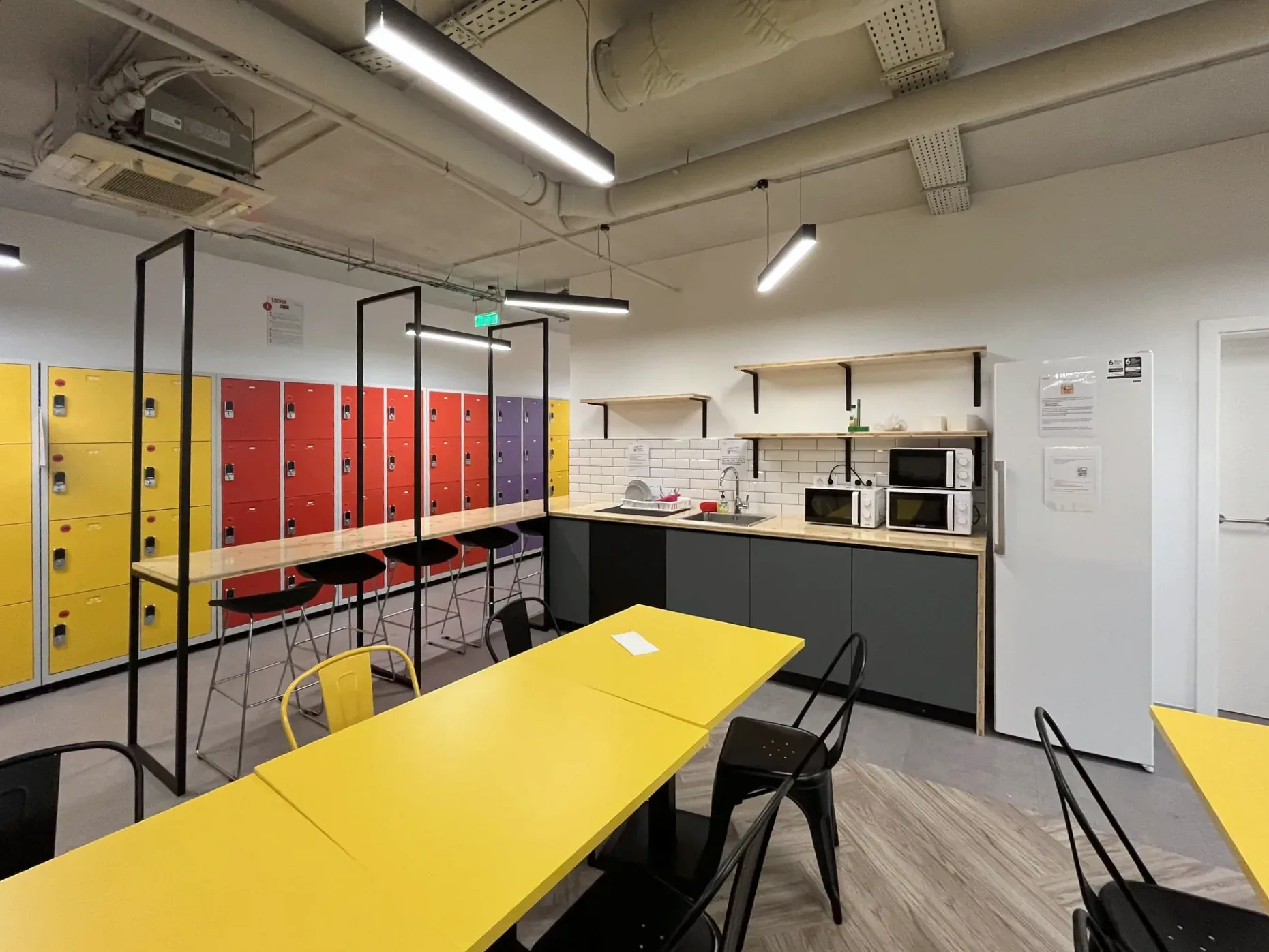 Mesas de tampo amarelo com cadeiras metálicas pretas em primeiro plano, a fila de cacifos coloridos atrás de pórticos de aço preto à esquerda, e uma copa ao fundo com azulejo branco tipo metro, prateleiras de madeira sobre cantoneiras pretas e móveis baixos cinzentos.