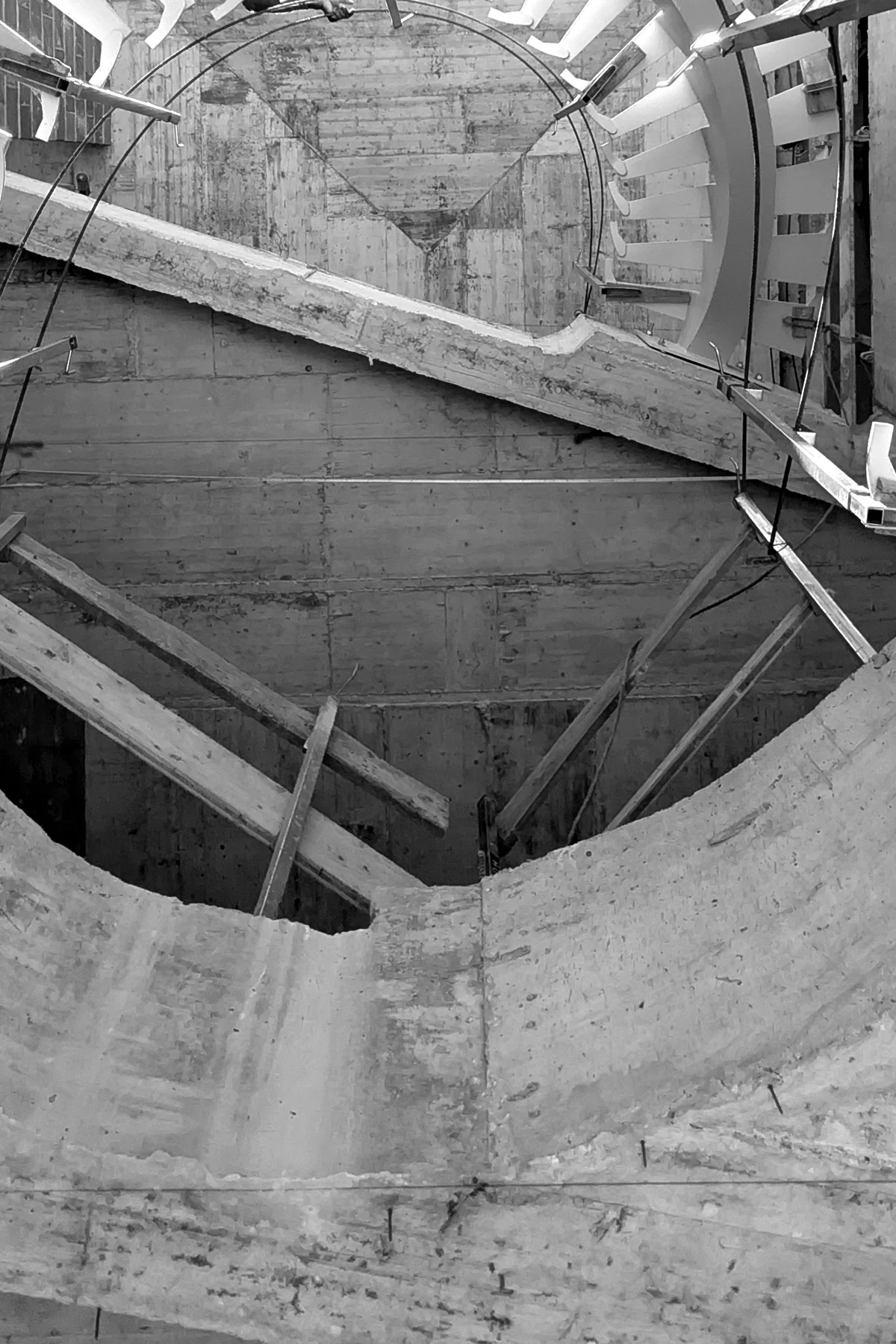 En blanco y negro, mirando hacia arriba dentro de un vacío circular: la cara inferior de una escalera helicoidal de hormigón que gira por encima, sus peldaños irradiando desde el muro, con piezas de encofrado y barras cruzando la abertura.