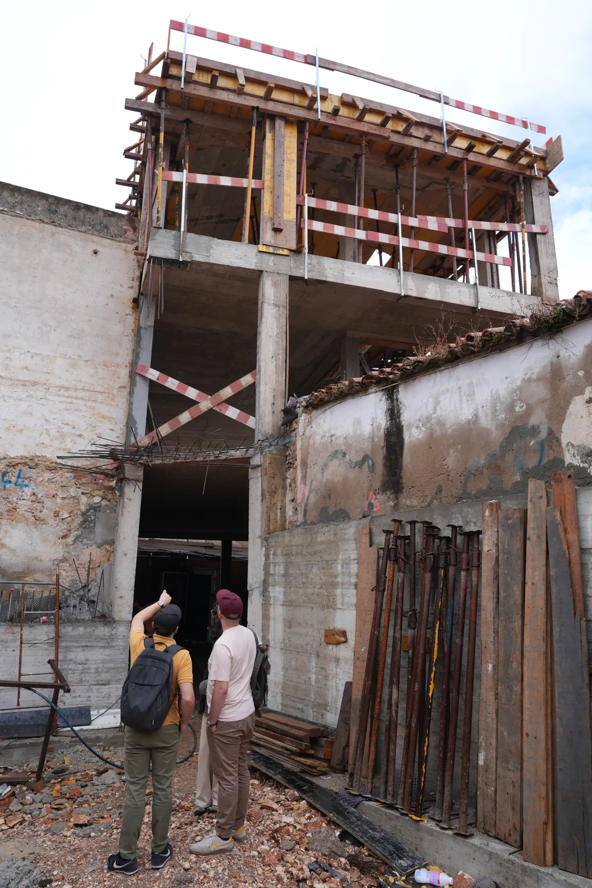 A street wall standing on its own, with a concrete structure rising behind and above it; two people at the foot of it looking up, and a set of uprights leaning against the wall.