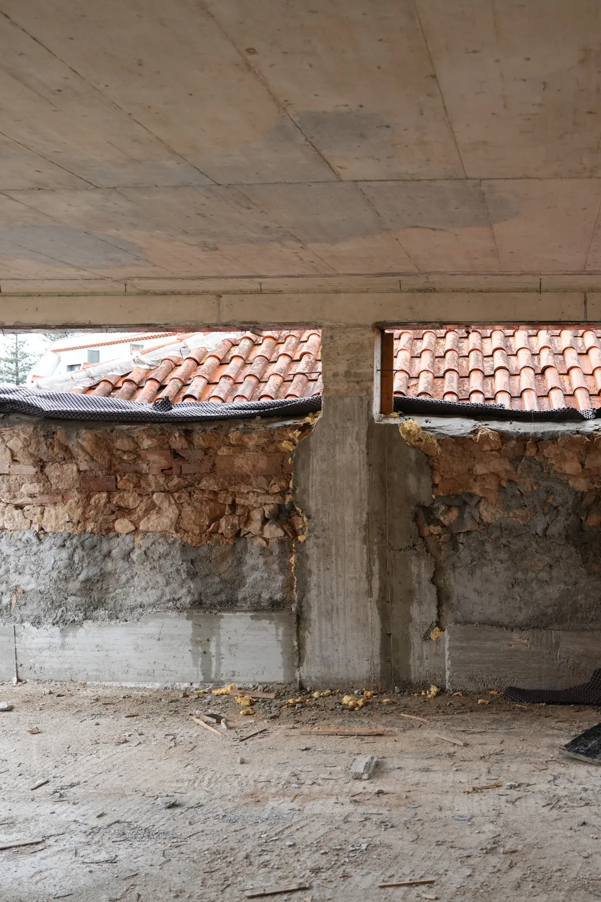 Inside the structure: a concrete soffit and vertical elements above, a stone masonry wall below, and two long openings through which neighbouring tiled roofs are visible.