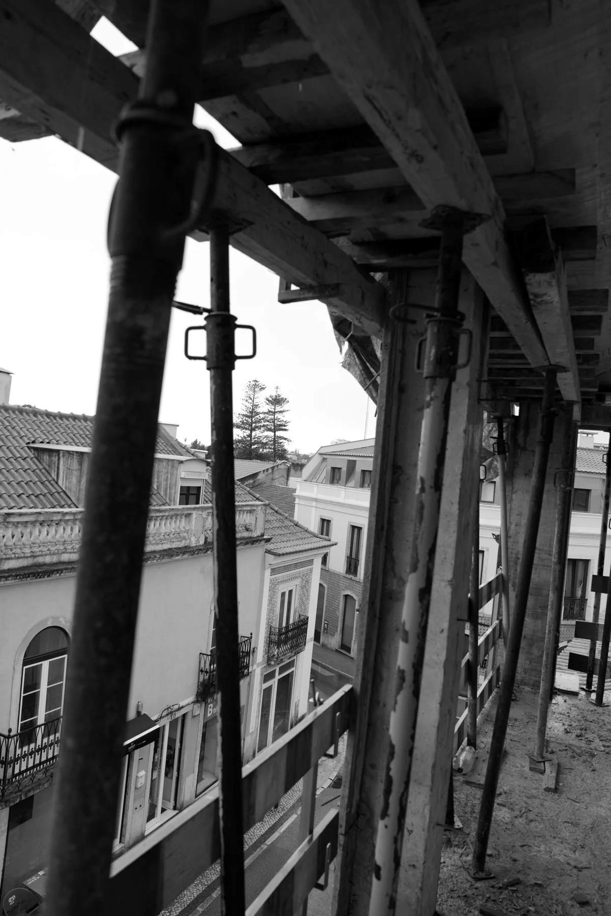 Black and white, looking out from the edge of a floor between uprights and boards to the balconied façades of the street below.