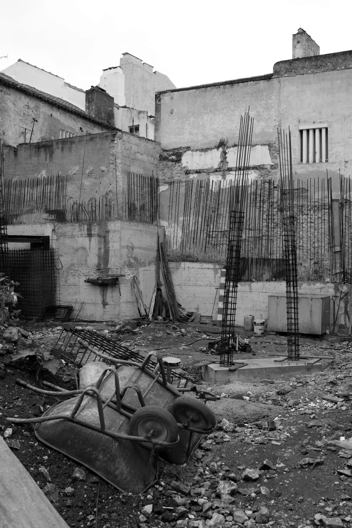Black and white: an area of the site between existing buildings, with walls all round, bars standing out of the foundations, rubble and a wheelbarrow on the ground.