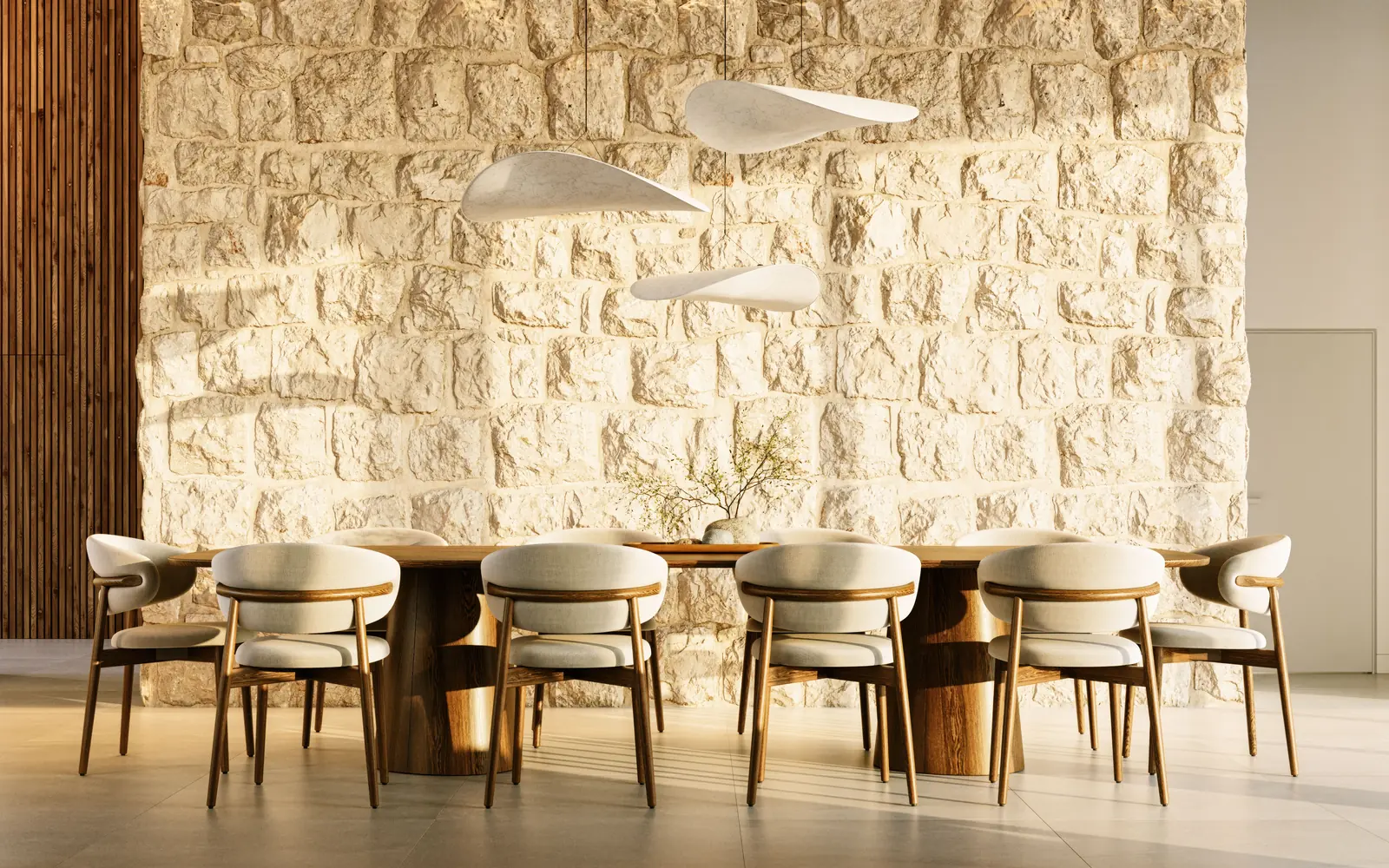 The same stone wall seen head on, lit from above, with a long table for ten and pale upholstered chairs against it.