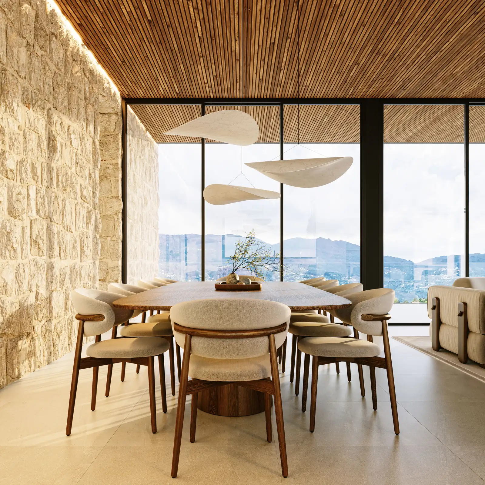 The dining room seen against a wall of dressed stone, a round table with six chairs, three pale leaf-shaped pendants above it and the valley through the glazing on the left.