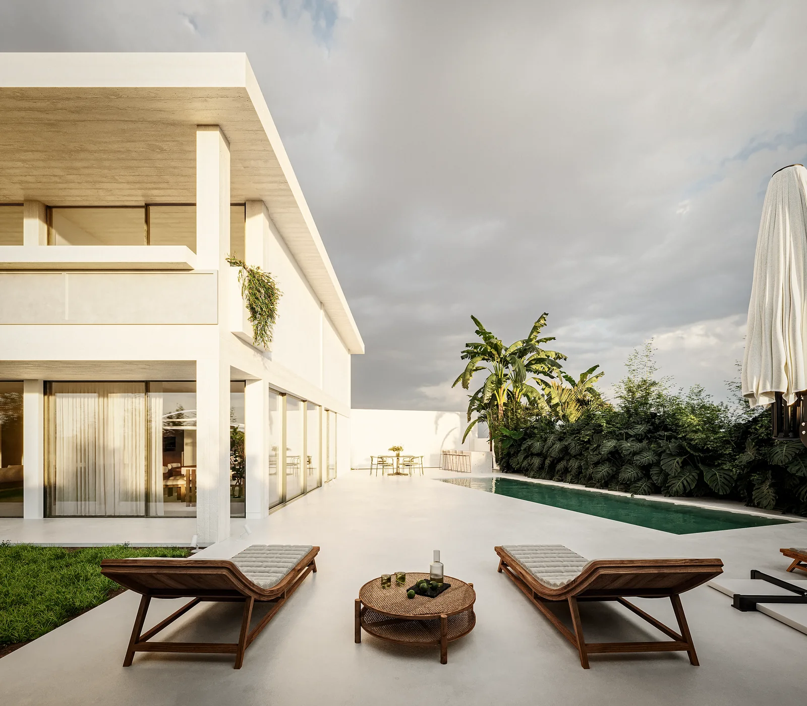 O terraço da piscina: um tanque rectangular escuro embutido em pavimento de pedra clara, duas espreguiçadeiras de madeira e uma mesa baixa redonda, a casa atrás com o piso térreo envidraçado aberto para o terraço.