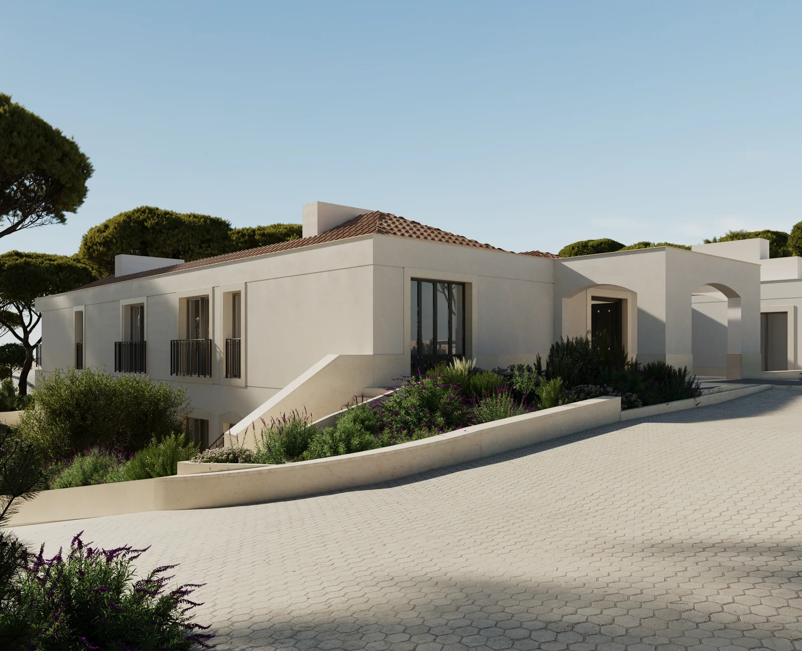 The arrival side: a paved forecourt curving up to a plain white elevation with tall shuttered openings, a low retaining wall of planting to one side and pines closing the horizon.